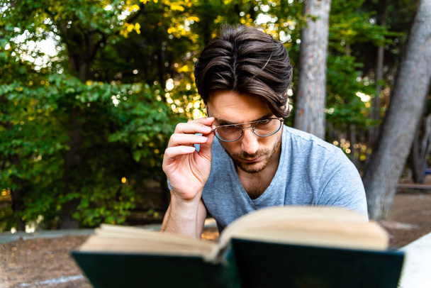 Estudante masculino com óculos lê cuidadosamente um livro antigo em um parque
 - Foto, Imagem