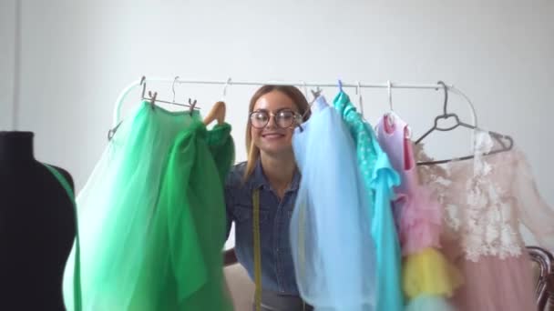 Beautiful blonde seamstress gleefully looks out from behind clothes on a hanger - Footage, Video