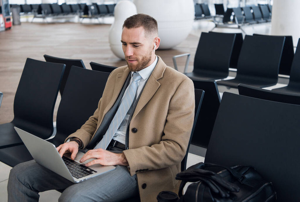 Always on the go. Portrait of handsome smiling businessman in formalwear holding a laptop with earphones while sitting in the airport - Fotó, kép