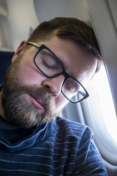 Νεαρός, γενειοφόρος άντρας με γυαλιά κοιμάται ήρεμα στο αεροπλάνο, ακουμπώντας το κεφάλι του στο παράθυρο.. - Φωτογραφία, εικόνα