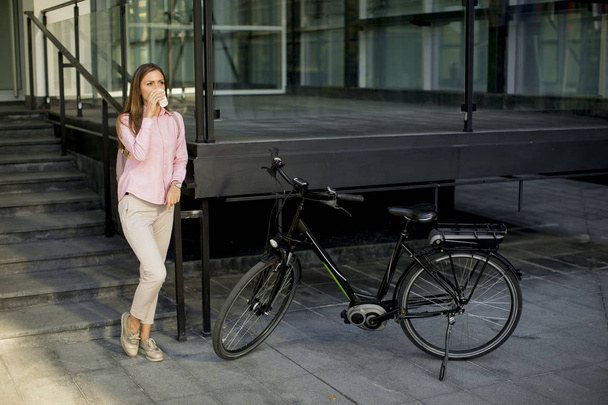 Belle jeune cycliste boit du café dans une tasse par le vélo électrique dans l'environnement urbain
 - Photo, image