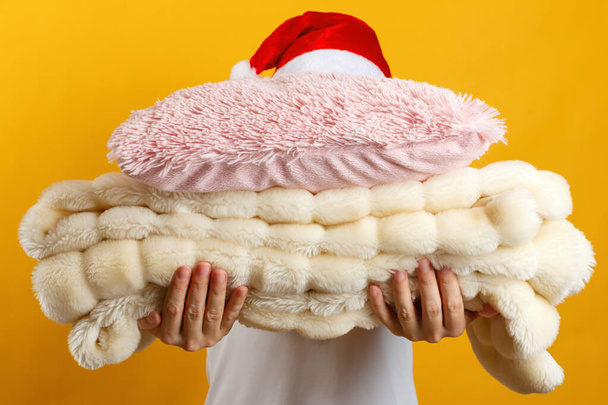Woman in santa hat holding a stack of warm fluffy plaid and pillows on a background of a yellow wall - Foto, Imagem