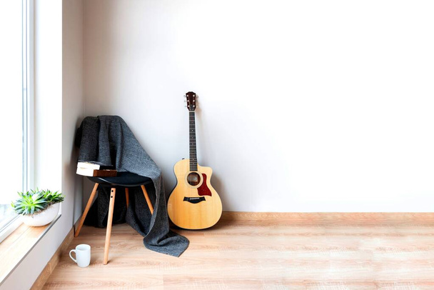 Interior de casa contemporânea. Cadeira preta coberta com cobertor cinza lanoso e guitarra acústica na frente de uma parede branca vazia. Tempo para hobby e lazer
. - Foto, Imagem