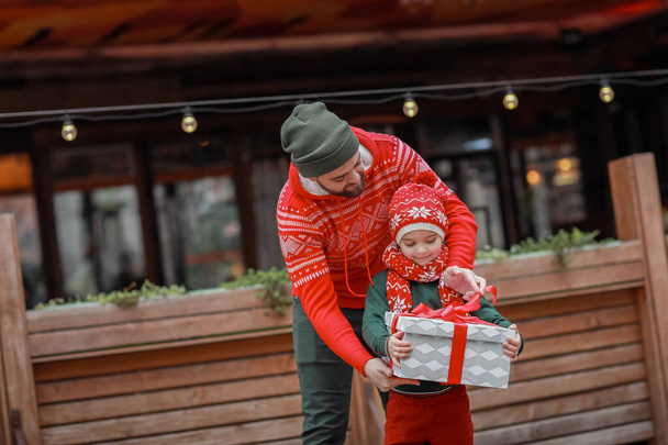 Dad with his son in bright holiday clothes posing in front of the camera on the street and holding a box with a gift in their hands. - Fotografie, Obrázek