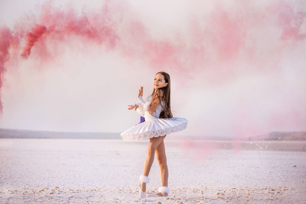 Tender young ballerina dancer in a snow-white tutu dress and white pointe shoes in pink smoke.  on a salty dried lake. Fantastic landscape and a girl  ballerina - Foto, Bild