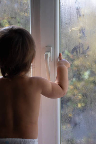 A small child looks out of a foggy window, points a finger at the street. Dangerous situation - the baby may open the door and fall from a height - Foto, afbeelding