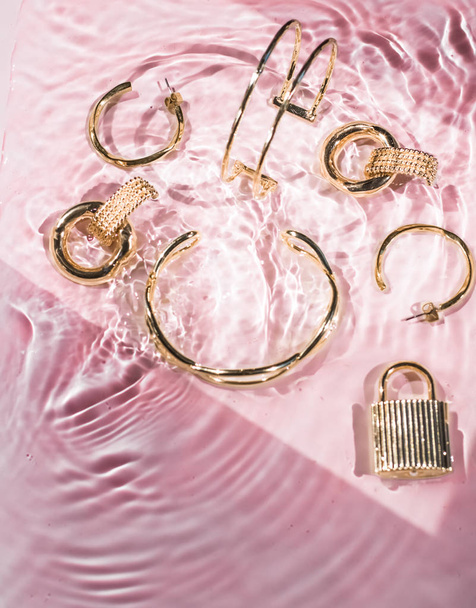 Złote bransoletki, kolczyki, pierścionki, biżuteria na różowej wody backgr - Zdjęcie, obraz
