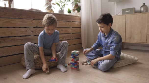 Two boys brothers are building a tower from wooden blocks sitting on the floor - Imágenes, Vídeo