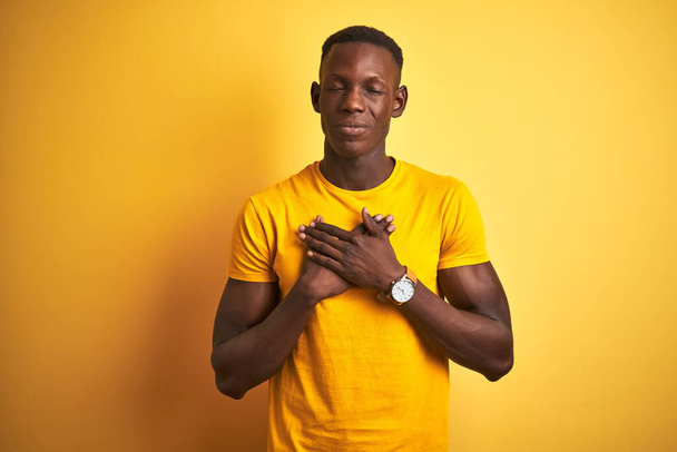 Fiatal afro-amerikai férfi alkalmi pólóban áll izolált sárga háttér felett mosolyogva, csukott szemmel és hálás gesztussal a mellkasán. Egészségügyi koncepció. - Fotó, kép