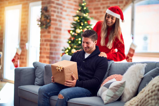 Young beautiful couple smiling happy and confident. Woman surprise man with gift  around christmas tree at home - Photo, Image