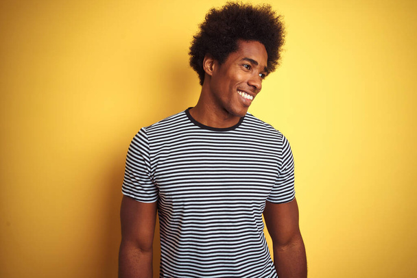 Африканський американець з волоссям афро, одягнений у військово-морську смугасту футболку над ізольованим жовтим фоном, який дивиться в сторону з посмішкою на обличчі, природним виразом. Сміючись упевнено. - Фото, зображення