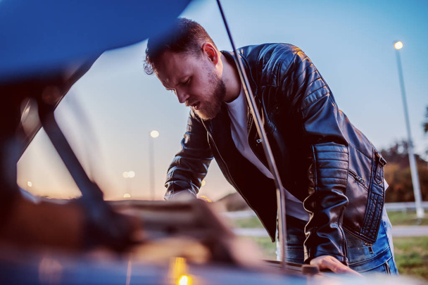 Young serious handsome caucasian bearded blond man in leather jacket looking engine of his car under the hood. Something went wrong. - Photo, Image