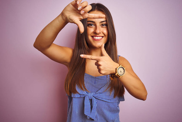 Young beautiful woman wearing striped dress standing over isolated pink background smiling making frame with hands and fingers with happy face. Creativity and photography concept. - Valokuva, kuva