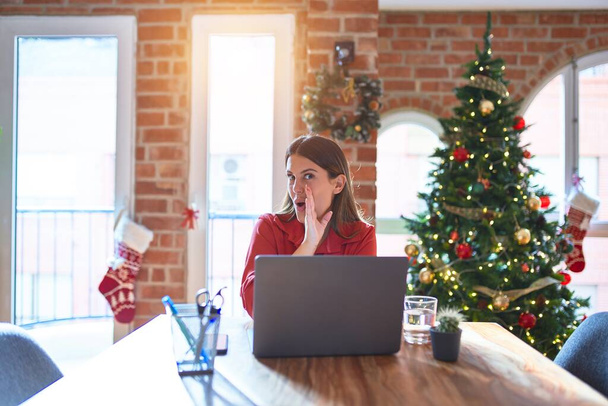 美しい女性ですテーブルで作業ノートパソコンと一緒に家の周りにクリスマスツリーの手口で秘密の噂を伝える,悪意のある話の会話をささやく - 写真・画像