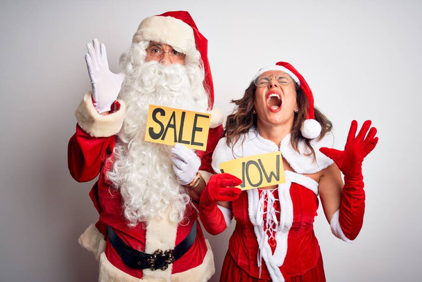Couple wearing Santa costume holding wow and sale banner over isolated white background crazy and mad shouting and yelling with aggressive expression and arms raised. Frustration concept. - Fotografie, Obrázek