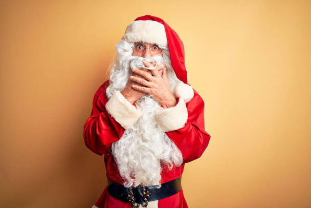 Ein gutaussehender Mann mittleren Alters im Weihnachtsmannkostüm, der vor isoliertem, gelbem Hintergrund steht, schockiert den Mund mit den Händen versehentlich bedeckt. Geheimes Konzept. - Foto, Bild