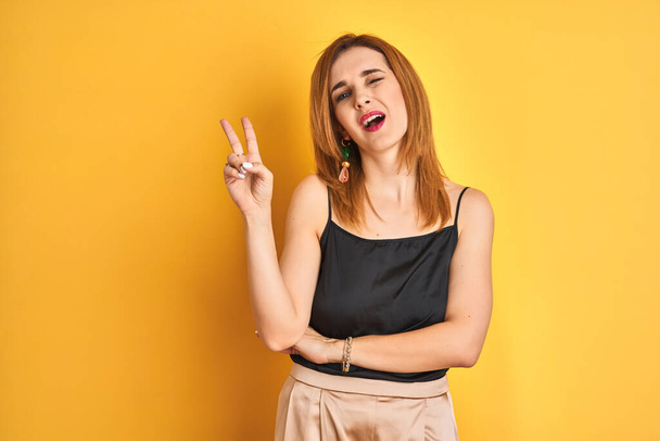 Redhead caucasian business woman standing over yellow isolated background smiling with happy face winking at the camera doing victory sign. Number two. - 写真・画像