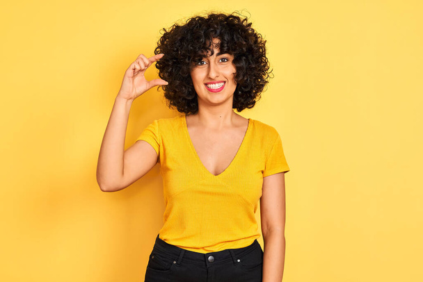 孤立した黄色の背景の上に立ってTシャツを着て巻き毛を持つ若いアラブの女性は笑顔と指を見てとカメラで小さなサイズの看板を行う手で自信を持ってジェスチャー。概念の測定. - 写真・画像
