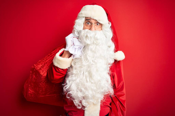 Keski-ikäinen mies yllään Santa puku tilalla säkki lahjoja yli eristetty punainen tausta peloissaan shokissa yllätys kasvot, peloissaan ja innoissaan pelko ilme
 - Valokuva, kuva