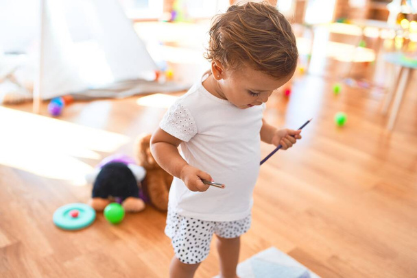 Чарівний малюк тримає олівці, що стоять навколо багатьох іграшок в дитячому садку
 - Фото, зображення