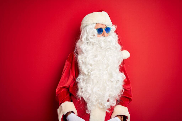 Muž středního věku v kostýmu Santa Clause a slunečních brýlích na izolovaném červeném pozadí, dívá se stranou s úsměvem na tváři, přirozený výraz. Smějící se sebevědomí. - Fotografie, Obrázek