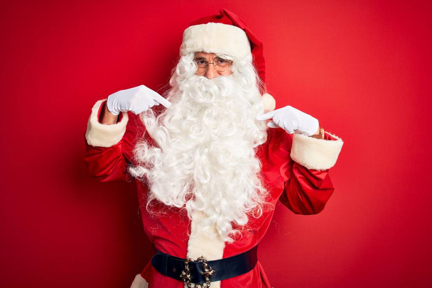 Hombre guapo de mediana edad vistiendo traje de Santa de pie sobre fondo rojo aislado mirando confiado con sonrisa en la cara, señalándose con los dedos orgullosos y felices
. - Foto, Imagen