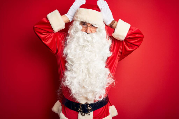 Pohledný muž středního věku v kostýmu Santa stojí nad izolovaným červeným pozadím a dělá gesto králičích uší dlaněmi, které vypadají cynicky a skepticky. Koncept velikonočního králíka. - Fotografie, Obrázek