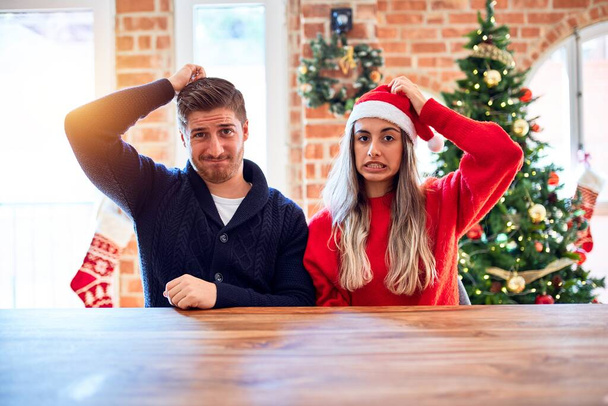 junges Paar mit Weihnachtsmannmütze, das zu Hause auf Stuhl und Tisch um den Weihnachtsbaum sitzt, verwirrt und wundert sich über die Frage. Unsicher im Zweifel, mit der Hand auf dem Kopf denkend. Nachdenkliches Konzept. - Foto, Bild