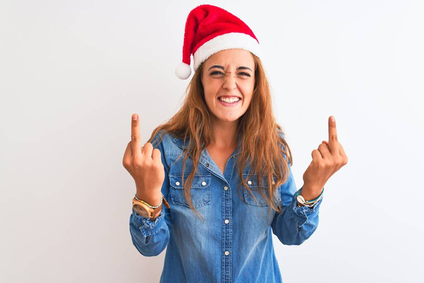 孤立した背景の上にクリスマスの帽子をかぶって若い美しい赤毛の女性あなたに悪い表情、挑発、失礼な態度をファックしている中指を示しています。興奮して悲鳴 - 写真・画像