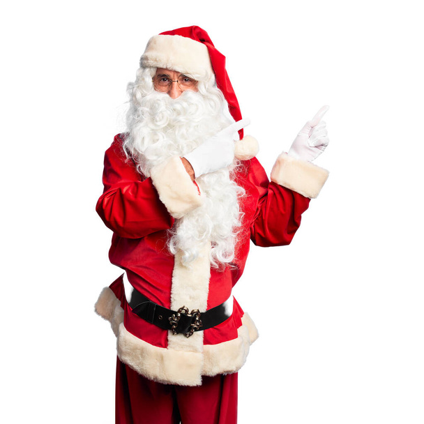 Homem bonito de meia idade vestindo traje de Papai Noel e barba de pé sorrindo e olhando para a câmera apontando com duas mãos e dedos para o lado
. - Foto, Imagem