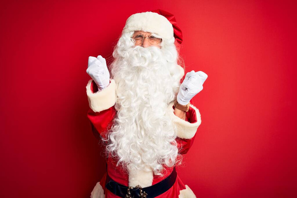 Ein gutaussehender Mann mittleren Alters im Weihnachtsmannkostüm steht vor isoliertem roten Hintergrund und feiert verrückt und verrückt nach Erfolg mit erhobenen Armen und geschlossenen Augen, die aufgeregt schreien. Siegerkonzept - Foto, Bild
