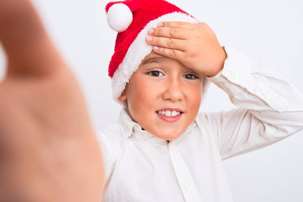 Menino bonito vestindo chapéu de Natal Santa fazer selfie sobre fundo branco isolado estressado com a mão na cabeça, chocado com vergonha e rosto surpresa, irritado e frustrado. Medo e chateado por erro
. - Foto, Imagem