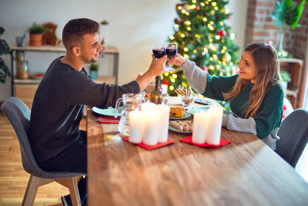 Jovem casal bonito sorrindo feliz e confiante. Comer alimentos e brindar com xícara de vinho celebrando o Natal em casa
 - Foto, Imagem