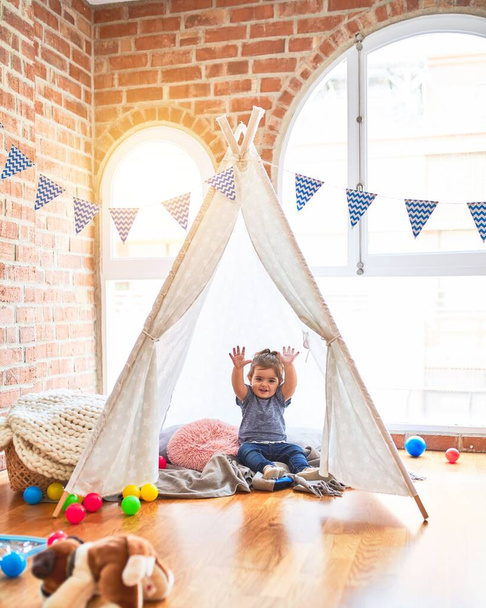 Beautiful toddler sitting on the floor inside tipi with hands raised smiling at kindergarten - Фото, зображення