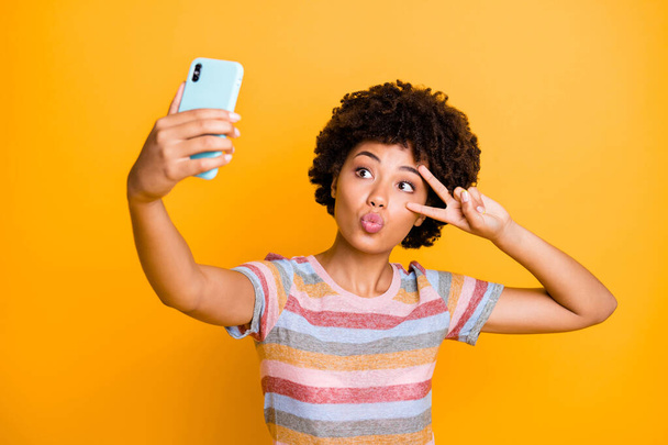 De cerca la foto de la romántica chica mulata soñadora blogger viaje tomar selfie en el teléfono inteligente para el blog hacer v-signos enviar besos de aire usar elegante camiseta a rayas aislado color amarillo fondo
 - Foto, imagen