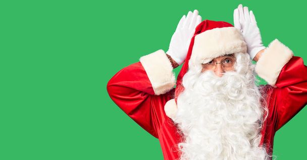 Ein gutaussehender Mann mittleren Alters im Weihnachtsmann-Kostüm und mit Bart steht da und macht Hasenohren-Gesten, während Handflächen zynisch und skeptisch aussehen. Osterhasen-Konzept. - Foto, Bild