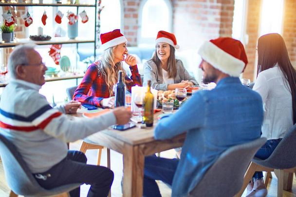 Hermosa familia vistiendo sombrero de santa claus reunión sonriente feliz y seguro. Comer pavo asado celebrando la Navidad en casa
 - Foto, Imagen