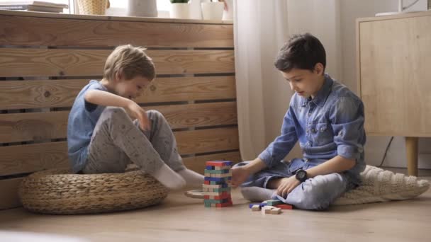 Two boys brothers are building a tower from wooden blocks sitting on the floor - Filmati, video