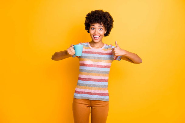 Excellent quality arabica. Portrait of cheerful amazed afro american girl hold mug beverage show thumb up advertise promo wear striped t-shirt brown pants trousers isolated yellow color background - Fotografie, Obrázek