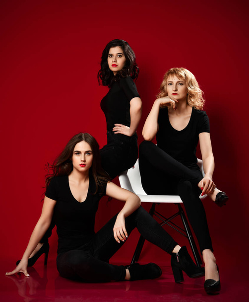Three young sexy women in black t-shirts and black tight jeans are sitting on at and near white chair on red - Фото, изображение