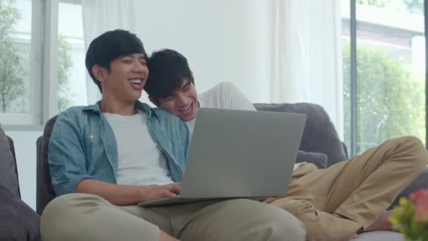 Young Gay couple using computer laptop at modern home. Asian LGBTQ men happy relax fun using technology watching movie in internet together while lying sofa in living room at house concept. - Video