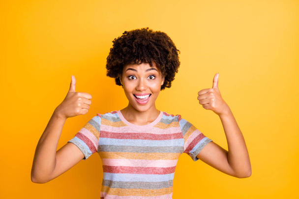 Photo of beautiful dark skin lady holding thumbs raised air expressing agreement and positive attitude wear casual striped t-shirt isolated yellow background - Foto, Bild