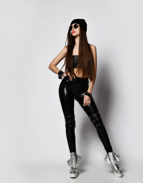 Brutal girl in round sunglasses, black hat, tight leather leggings with zippers is cool posing looking up st copy space - Photo, Image