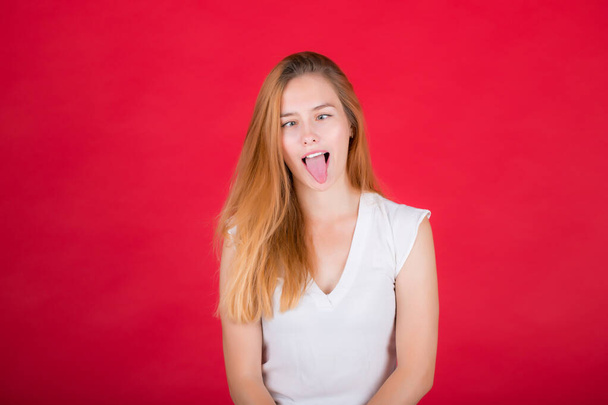 A young woman is grimacing at the camera. Girl in a white shirt on a red background shows tongue. - Fotó, kép