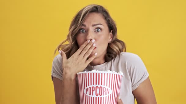 Focused young woman with red lips holding a basket of popcorn and showing disgusting while looking at the camera over yellow background isolated - Felvétel, videó