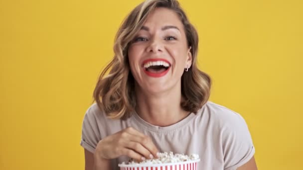 Happy young woman with red lips holding a basket of popcorn watching a comedy and laughing over yellow background isolated - Video