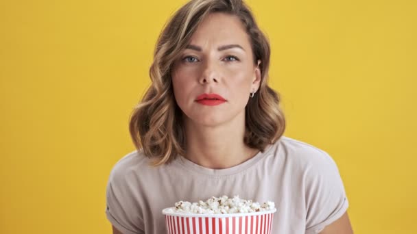 Focused on wathing a movie young woman with red lips holding a basket of popcorn and showing disgusting and feeling scary while looking at the camera over yellow background isolated - Кадры, видео