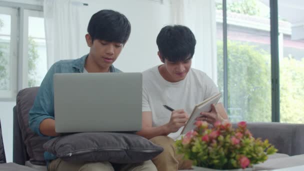 Young Asian Gay couple working laptop at modern home. Asia LGBTQ+ men happy relax fun using computer and analyzing their finances in internet together while lying sofa in living room at house concept. - Video