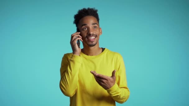 Smiling african american man in yellow wear talking on mobile phone over blue background. Handsome man holding and using smart phone. Guy with technology - 映像、動画