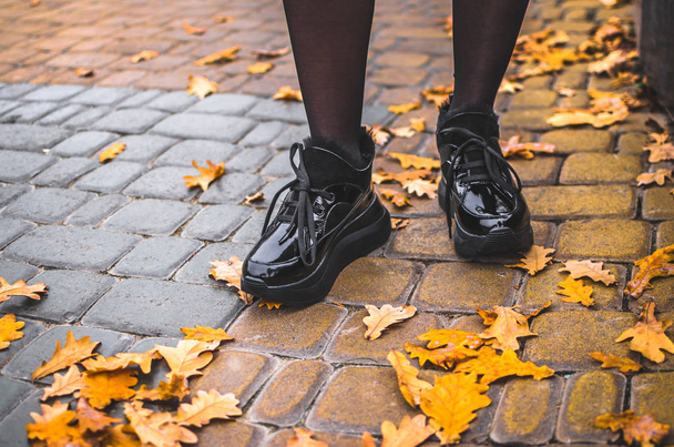 Ένα κορίτσι στέκεται σε μαύρο λαμπερό ζεστά παπούτσια σε ένα κεραμίδι με φύλλα στο κέντρο της πόλης σε ένα πάρκο - Φωτογραφία, εικόνα